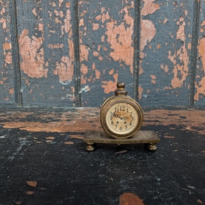 Peut inclure: Une petite horloge de bureau ronde en laiton avec un cadran crème, des chiffres arabes noirs et deux aiguilles sombres. L'horloge est fixée sur une base rectangulaire en laiton dotée de quatre petits pieds, offrant un aspect métallique vieilli.