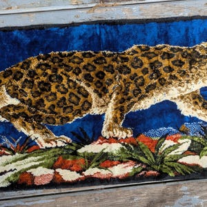 Puede incluir: Alfombra rectangular con un diseño detallado de leopardo. El leopardo, con pelaje marrón y manchas marrones, se encuentra sobre un fondo azul. La alfombra tiene flecos azules en los bordes. La alfombra probablemente está hecha de tela.