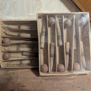 May include: Two vintage sets of what appear to be antique cutlery or serving pieces. Each set is in a cardboard box with individual slots for the items. The pieces are light-colored with dark accents, and the boxes are sealed with clear plastic.