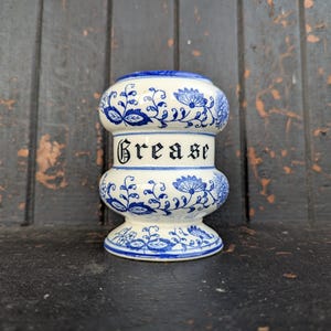 Puede incluir: Un tarro de grasa de cerámica blanca y azul con motivos florales y la palabra "Grease" en letras negras. El tarro tiene forma bulbosa con tapa y base, y está decorado con diseños florales azules.