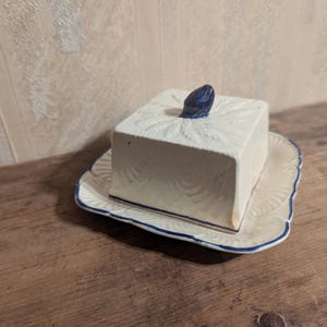 Può includere: Una burriera in ceramica bianca con coperchio quadrato e pomello blu. Il piatto poggia su un piatto abbinato con bordo smerlato e rifiniture blu. La burriera è su una superficie di legno.