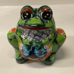 Może przedstawiać: Zielona ceramiczna figurka żaby z kolorowymi wzorami kwiatowymi i czarnymi oczami. Żaba siedzi z nogami schowanymi pod ciałem.