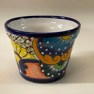 Puede incluir: Una maceta de cerámica colorida con un borde azul y un interior blanco. La maceta está decorada con un patrón floral en tonos de amarillo, naranja, verde, azul y rojo.