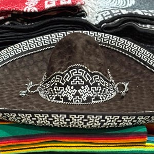Puede incluir: Un sombrero marrón con ala ancha y un patrón geométrico decorativo blanco y negro. El sombrero presenta una corona detallada y una correa para la barbilla. El sombrero se exhibe encima de una pila de mantas coloridas.