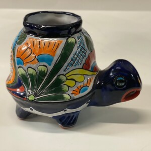 May include: A hand-painted ceramic turtle planter with a blue base and colourful floral designs. The turtle has a small opening on its back for planting.