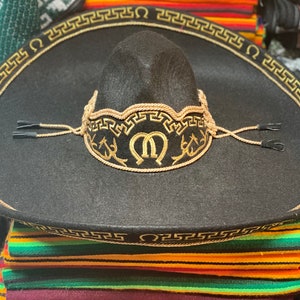Puede incluir: Un sombrero negro con bordados dorados que representan un diseño de herradura y un patrón de llave griega alrededor del borde. El sombrero está sentado encima de una pila de mantas coloridas.