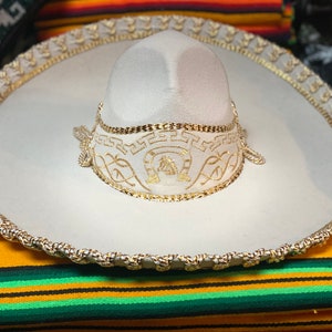Op de afbeelding: Een witte sombrero met gouden randen en borduursels. De hoed heeft een afgeronde kroon en een brede, platte rand. De rand en de kroon zijn versierd met gouden touwranden en decoratieve borduursels. De hoed ligt op een kleurrijk gestreept textiel.