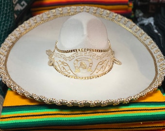 May include: A white sombrero with gold trim and embroidery. The hat features a rounded crown and a wide, flat brim. The brim and crown are adorned with gold rope trim and decorative embroidery. The hat is resting on a colorful striped textile.