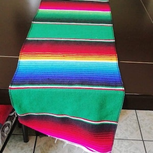 May include: A colorful Mexican table runner with green, red, yellow, blue, and white stripes. The runner is made of woven fabric and has a fringed edge.