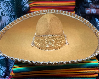 May include: A brown sombrero with gold embroidery of a horseshoe and a decorative trim. The sombrero is sitting on top of a colorful striped blanket.