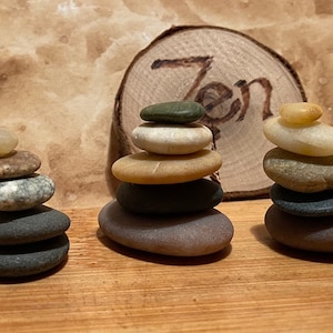 May include: Three stacks of smooth, gray, brown, and white stones are balanced on a wooden surface. The word "Zen" is carved into a wooden disc behind the stacks.