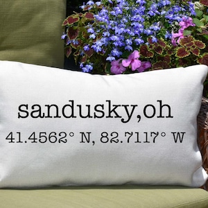 May include: White cushion with black text that reads "sandusky, oh 41.4562° N, 82.7117° W".