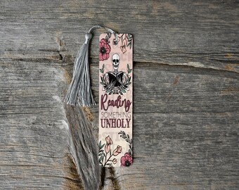 Può includere: Un segnalibro rosa e bianco con uno scheletro che legge un libro. Il segnalibro ha un design floreale e il testo "Reading something unholy".
