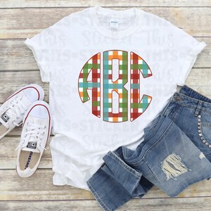 Puede incluir: Una camiseta blanca con un diseño de monograma redondo de cuadros de colores con las letras "ABC". La camiseta está colocada sobre una superficie de madera con un par de zapatillas blancas y unos vaqueros azules.