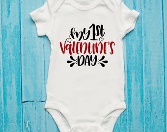 May include: White baby onesie with the text "my 1st Valentine's Day" in black and red. The onesie has short sleeves and a snap closure. The text includes heart accents. The onesie is displayed on a blue wooden surface.