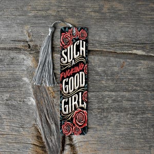 May include: A decorative bookmark with a gray tassel. The bookmark features the text "SUCH A FUCKING GOOD GIRL" in a stylized font, with red roses and a black background. The bookmark is on a wooden surface.