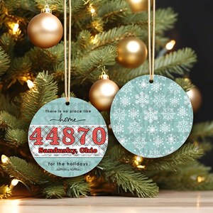 Op de afbeelding: Een ronde kerstbal met een blauw en wit sneeuwvlokkenpatroon. De andere kant van de kerstbal heeft een rood-wit geruite achtergrond met de tekst "There is no place like home 44870 Sandusky, Ohio for the holidays."