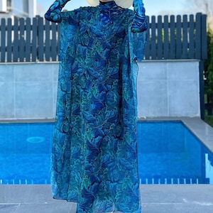 Puede incluir: Un traje de ba&ntilde;o de manga larga con estampado floral azul y verde con una larga bata transparente a juego. El traje de ba&ntilde;o tiene cuello alto y un dise&ntilde;o modesto. La modelo lleva un sombrero de paja y sandalias blancas.