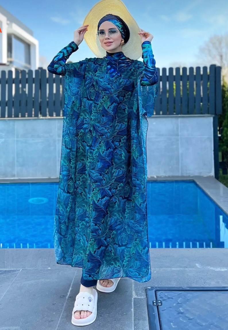 Puede incluir: Un traje de ba&ntilde;o de manga larga con estampado floral azul y verde con una larga bata transparente a juego. El traje de ba&ntilde;o tiene cuello alto y un dise&ntilde;o modesto. La modelo lleva un sombrero de paja y sandalias blancas.
