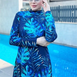 Puede incluir: Un traje de ba&ntilde;o azul con estampado de hojas tropicales. El traje de ba&ntilde;o tiene mangas largas y un cuello alto. La modelo lleva un pa&ntilde;uelo azul en la cabeza.