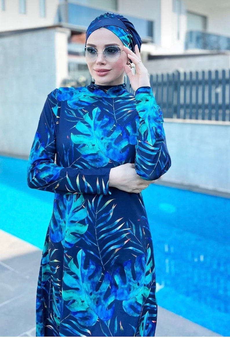 Puede incluir: Un traje de ba&ntilde;o azul con estampado de hojas tropicales. El traje de ba&ntilde;o tiene mangas largas y un cuello alto. La modelo lleva un pa&ntilde;uelo azul en la cabeza.