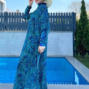 Puede incluir: Una mujer lleva un traje de ba&ntilde;o de manga larga con estampado floral azul y verde con un pa&ntilde;uelo a juego. Lleva un sombrero de paja y gafas de sol y est&aacute; de pie junto a una piscina.