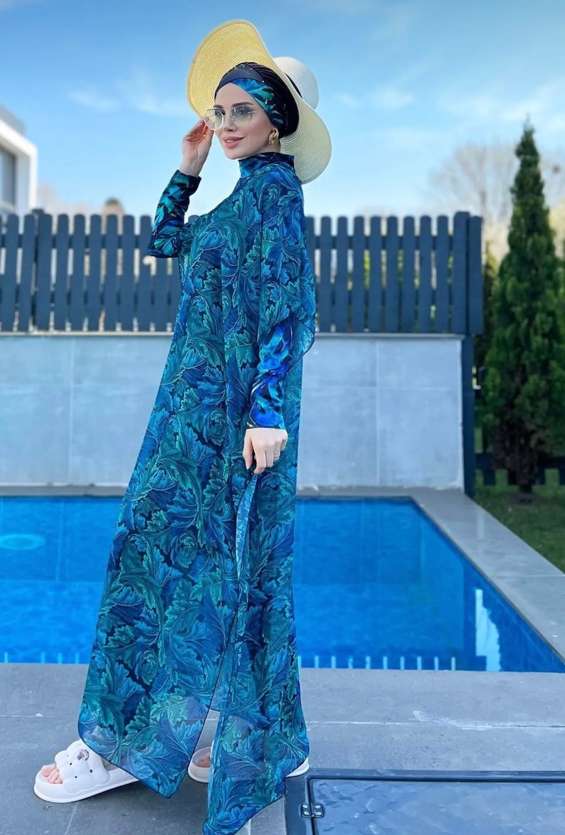Puede incluir: Una mujer lleva un traje de ba&ntilde;o de manga larga con estampado floral azul y verde con un pa&ntilde;uelo a juego. Lleva un sombrero de paja y gafas de sol y est&aacute; de pie junto a una piscina.