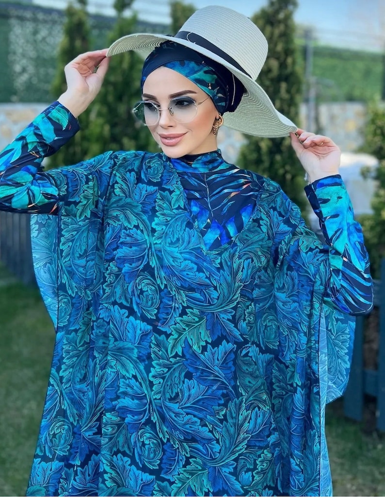 Puede incluir: Un traje de ba&ntilde;o de manga larga y cuello alto con estampado floral azul y verde. El traje de ba&ntilde;o tiene una superposici&oacute;n transparente con un estampado similar. La modelo lleva un sombrero de sol blanco con una banda negra.