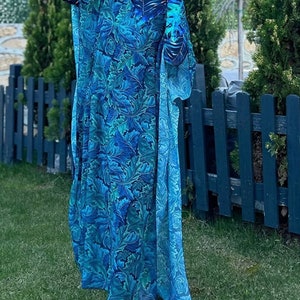 Puede incluir: Un traje de ba&ntilde;o de manga larga con estampado azul y verde con una bata a juego larga y fluida. El traje de ba&ntilde;o tiene cuello alto y la bata tiene un escote amplio y abierto. La modelo lleva un sombrero blanco y gafas de sol.