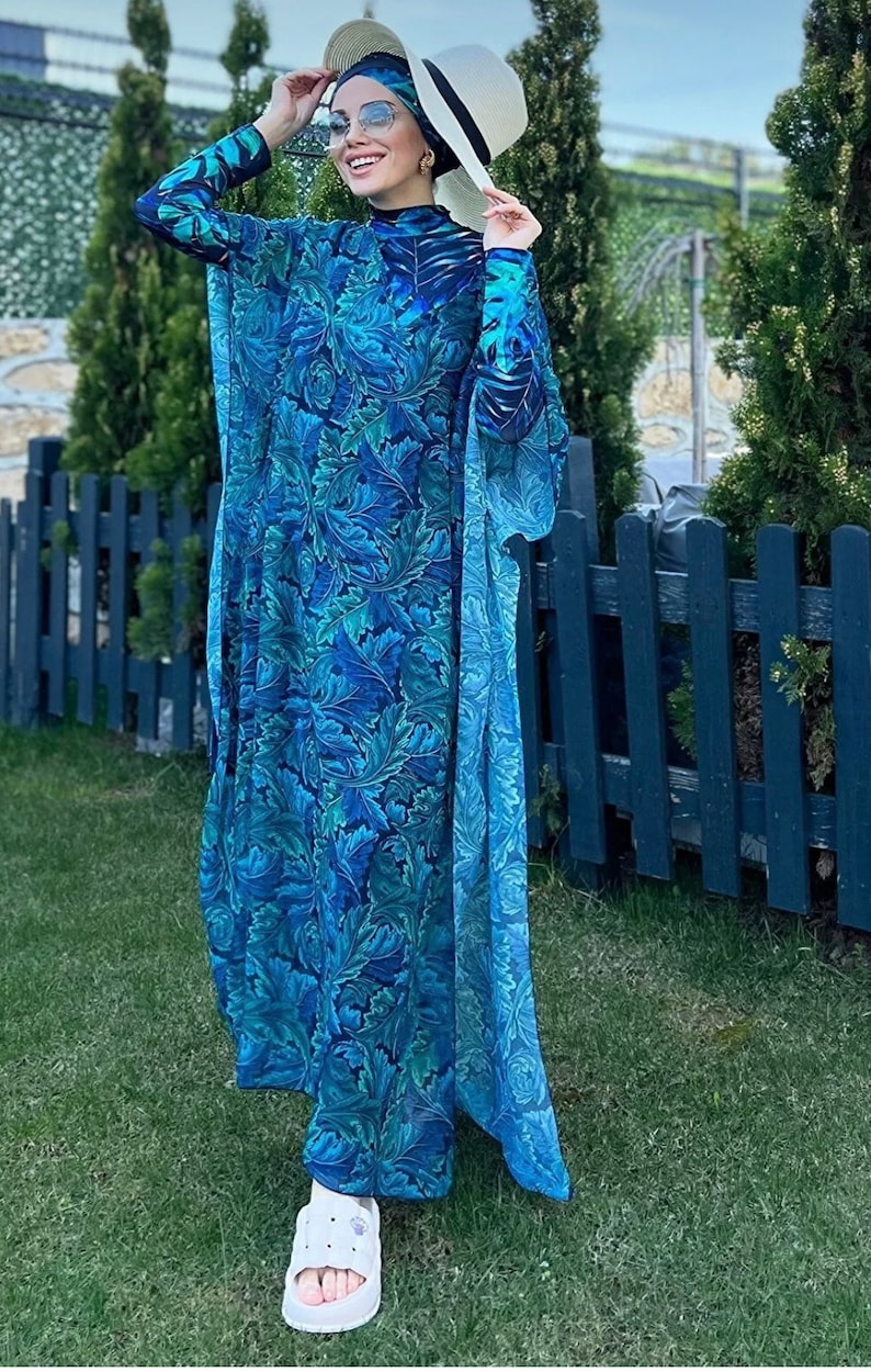 Puede incluir: Un traje de ba&ntilde;o de manga larga con estampado azul y verde con una bata a juego larga y fluida. El traje de ba&ntilde;o tiene cuello alto y la bata tiene un escote amplio y abierto. La modelo lleva un sombrero blanco y gafas de sol.