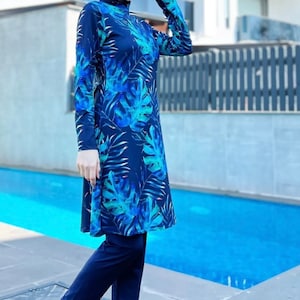 Puede incluir: Un traje de ba&ntilde;o azul y verde con estampado de hojas tropicales. El traje de ba&ntilde;o tiene mangas largas y pantalones. La modelo lleva un pa&ntilde;uelo azul y sandalias rojas.