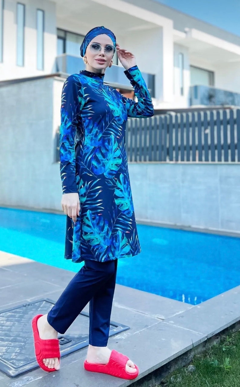 Puede incluir: Un traje de ba&ntilde;o azul y verde con estampado de hojas tropicales. El traje de ba&ntilde;o tiene mangas largas y pantalones. La modelo lleva un pa&ntilde;uelo azul y sandalias rojas.