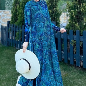 Puede incluir: Una mujer lleva un traje de ba&ntilde;o de manga larga con estampado floral azul y verde con un pa&ntilde;uelo a juego. Lleva un sombrero de sol blanco y sandalias blancas.