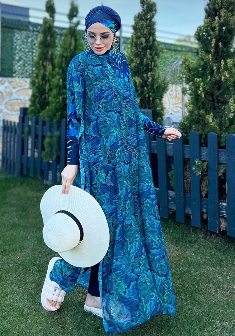 Puede incluir: Una mujer lleva un traje de ba&ntilde;o de manga larga con estampado floral azul y verde con un pa&ntilde;uelo a juego. Lleva un sombrero de sol blanco y sandalias blancas.