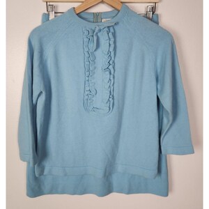May include: A light blue knit sweater and skirt set. The sweater has a round neckline, three-quarter sleeves, and a ruffled detail down the front. The skirt is a matching light blue color. The set is displayed on a wooden hanger.