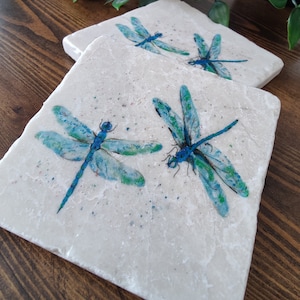 Pode incluir: Duas bases quadradas de pedra com desenhos de libélulas. As libélulas são pintadas em tons de azul e verde, com asas detalhadas. As bases têm um fundo salpicado esbranquiçado e estão colocadas sobre uma superfície de madeira.