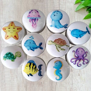 Puede incluir: Diez pomos redondos de cerámica con varias criaturas marinas pintadas en ellos. Los pomos presentan una estrella de mar, una ballena, una tortuga, una medusa, una foca, un narval, un pulpo, un pez, un caballito de mar y una tortuga.