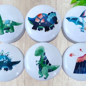 Może przedstawiać: Sześć białych, okrągłych gałek ceramicznych z kolorowymi ilustracjami dinozaurów. Gałki przedstawiają zielonego brachiozaura, niebieskiego stegozaura, zielonego T-Rexa, niebieskiego triceratopsa, niebieskiego pterodaktyla i czarny wulkan z czerwoną lawą.