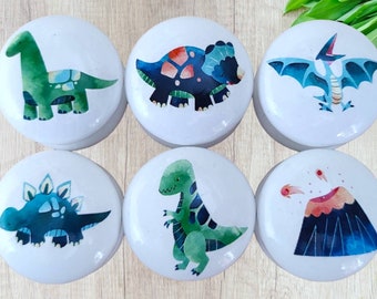 May include: Six white round ceramic knobs with colourful dinosaur illustrations. The knobs feature a green brachiosaurus, a blue stegosaurus, a green T-Rex, a blue triceratops, a blue pterodactyl, and a black volcano with red lava.