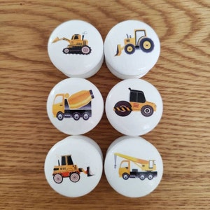 May include: Six white round knobs featuring various construction vehicle designs. The vehicles are yellow with black details. The designs include a bulldozer, tractor, cement mixer, steamroller, front loader, and crane.