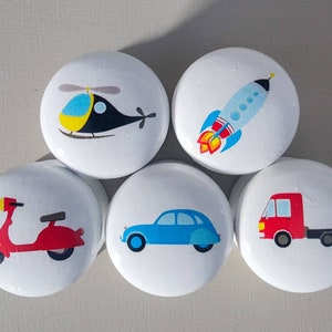 May include: Set of five white round knobs with colourful cartoon transportation designs. The designs include a red scooter, a blue car, a red truck, a blue and yellow helicopter, and a red and blue rocket.