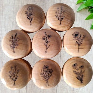 Może przedstawiać: Kolekcja ośmiu drewnianych gałek do szafek. Każda gałka ma inny czarny kwiatowy wzór, w tym maki, słoneczniki i inne dzikie kwiaty. Gałki są okrągłe i wykonane z jasnego drewna.