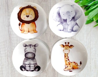 Puede incluir: Cuatro pomos redondos de cerámica con ilustraciones de acuarela de un león, un elefante, una cebra y una jirafa. Los pomos son blancos con un fondo marrón claro.