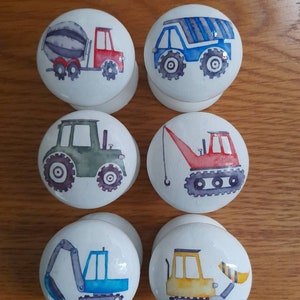 May include: Six white round knobs with different construction vehicle designs. The knobs feature a cement mixer truck, a dump truck, a tractor, a crane, an excavator, and a bulldozer.