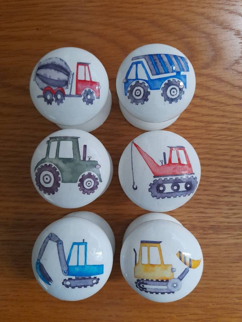 May include: Six white round knobs with different construction vehicle designs. The knobs feature a cement mixer truck, a dump truck, a tractor, a crane, an excavator, and a bulldozer.