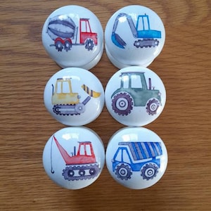 May include: Six white ceramic knobs with different construction vehicle designs. The knobs feature a cement mixer, excavator, bulldozer, tractor, crane, and dump truck.