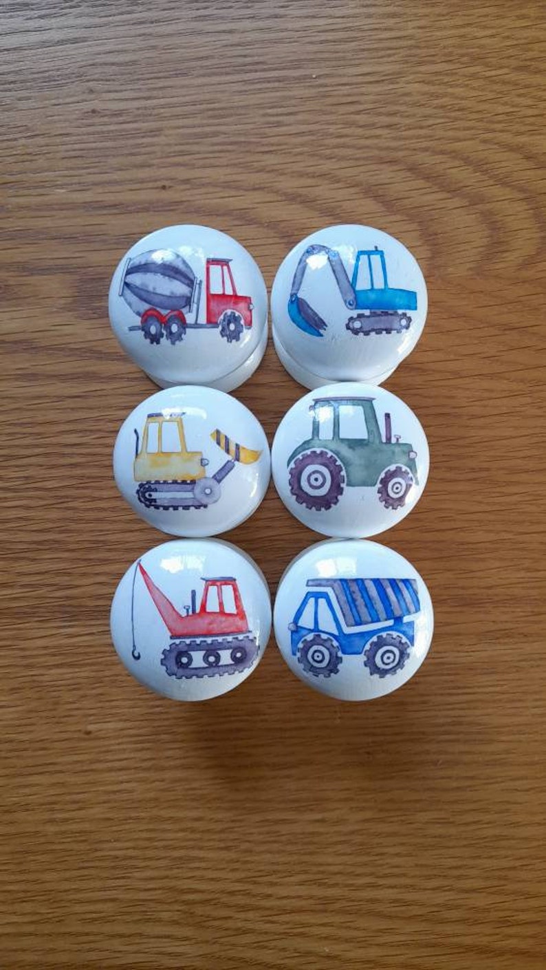 May include: Six white ceramic knobs with different construction vehicle designs. The knobs feature a cement mixer, excavator, bulldozer, tractor, crane, and dump truck.