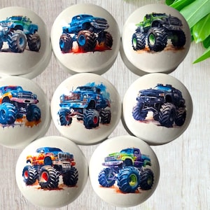 May include: Seven white ceramic cabinet knobs, each with a watercolour illustration of a monster truck. The trucks are in various colours, including blue, green, and orange, with large tyres.