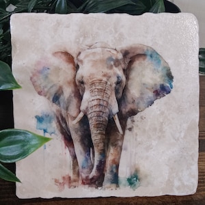 Op de afbeelding: Een aquarel schilderij van een olifant op een beige stenen onderzetter. De olifant kijkt naar voren met zijn slurf naar beneden. Het schilderij heeft een aquareleffect met spatten in blauw, groen en paars.