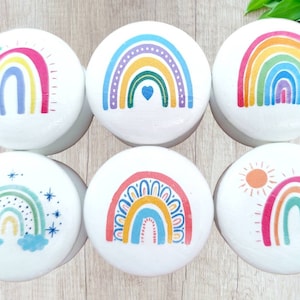 May include: Six white round wooden boxes with different rainbow designs. Each box features a rainbow with different colours and patterns, including stars, hearts, and a sun.